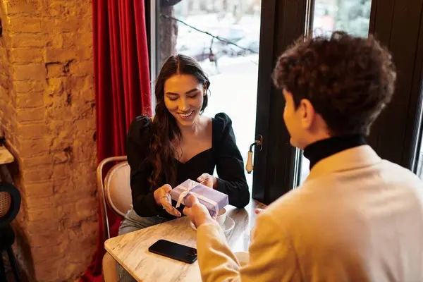 A young couple shares a heartfelt moment over dinner, exchanging gifts in a cozy restaurant setting. — Stock Photo