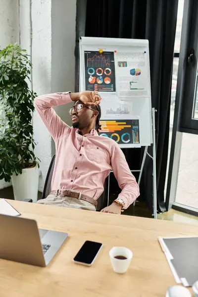 A young african american man relaxes while working remotely at a modern desk with charts behind him. — Stock Photo