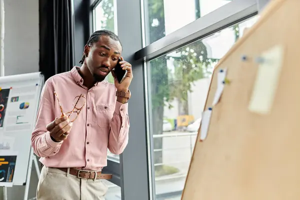 A young african american man speaks on his phone in a bright, stylish workspace while holding glasses. — Stock Photo