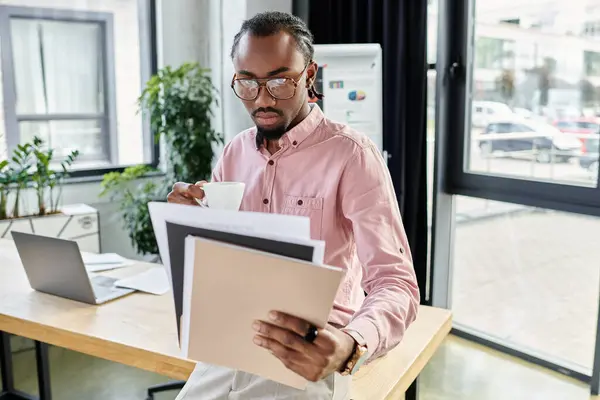 Young african american man sips coffee while reviewing documents in a bright workspace. — Stock Photo