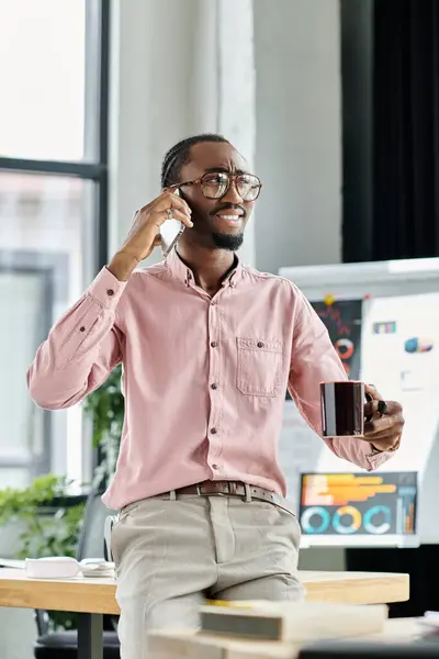 Cheerful digital nomad engages in a phone call with coffee in hand at a bright office setting. — Stock Photo
