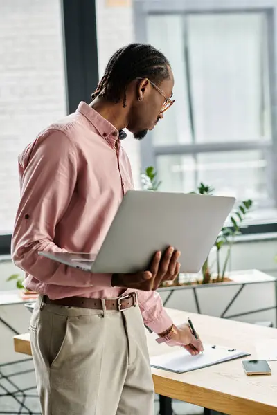 A young man is focused on his work, using a laptop and jotting notes in a vibrant office space. — Stock Photo