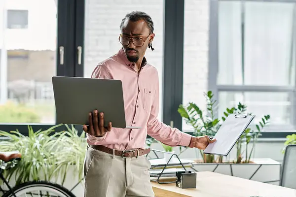 Focused young man uses laptop and clipboard in a stylish workspace filled with greenery. — Stock Photo