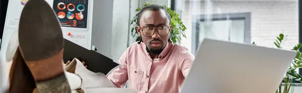 A young african american man works intently at his laptop in a dynamic and creative workspace. — Stock Photo