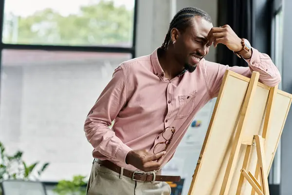 A young african american man stands by an easel, expressing creativity and focus in his workspace. — Stock Photo