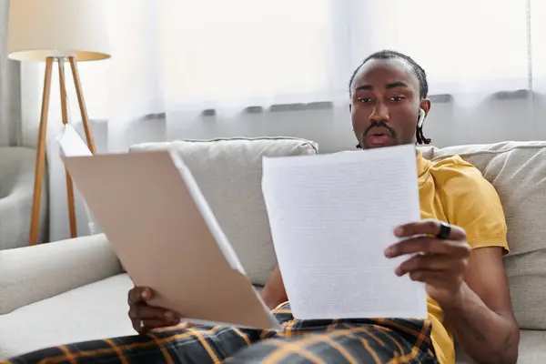 A young African American man reviews important documents on his couch while working remotely. — Stock Photo