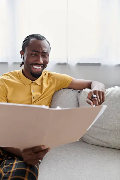 A young African American man sits comfortably on a couch, smiling as he reviews important documents. — Stock Photo