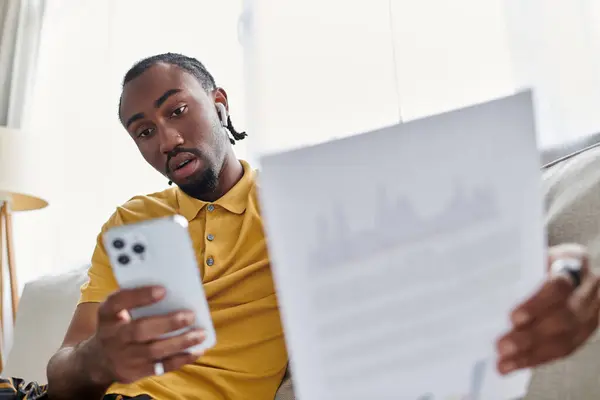 A young african american man reviews documents on his phone while relaxing indoors. — Stock Photo