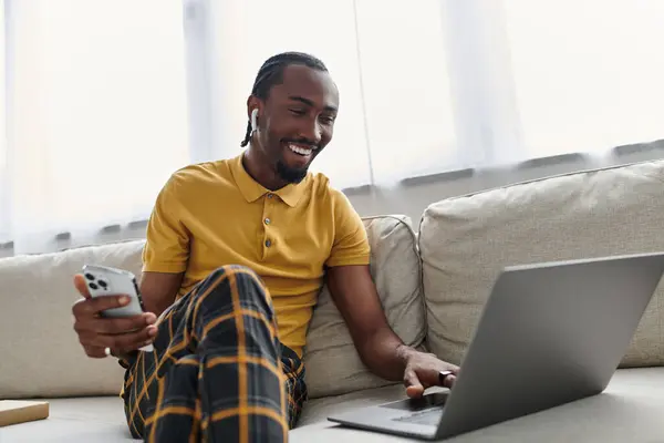 Participando en el trabajo remoto, un joven afroamericano sonríe mientras usa su computadora portátil y su teléfono inteligente. - foto de stock