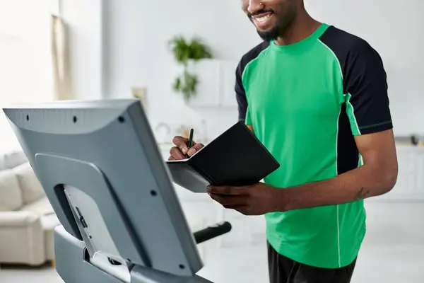 A young african american man smiles while writing notes during a workout session in a bright room. — Stock Photo