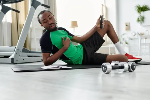 A young man on a mat uses his phone, surrounded by exercise gear in his fitness routine. — Stock Photo
