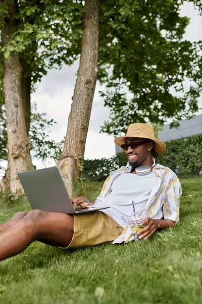 A stylish young man works on his laptop while relaxing on the grass under the shade of trees. — Stock Photo