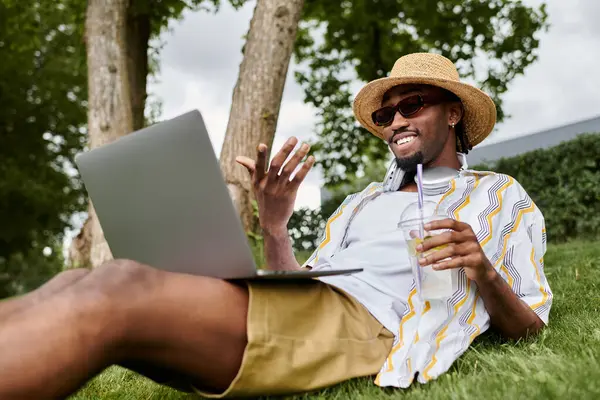 Un giovane uomo immerso nel lavoro sul suo portatile mentre si rilassa sull'erba all'aperto, sorseggiando un drink. — Foto stock