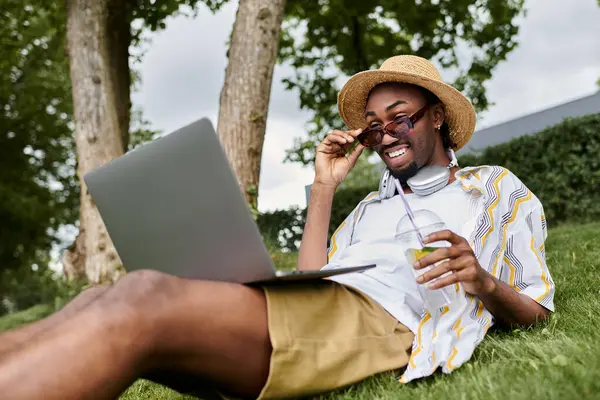 Digital nomad relaxes in a grassy park, smiling while engaging with his laptop and drink. — Stock Photo