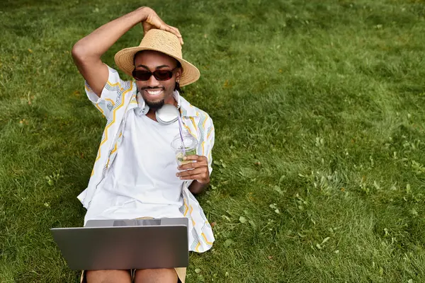 Smiling young man relaxes on grass, working on a laptop and enjoying a cool beverage in nature. — Stock Photo