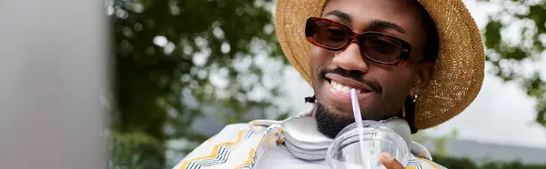 Young African American man smiles while sipping a cool beverage, immersed in remote work outdoors. — Stock Photo