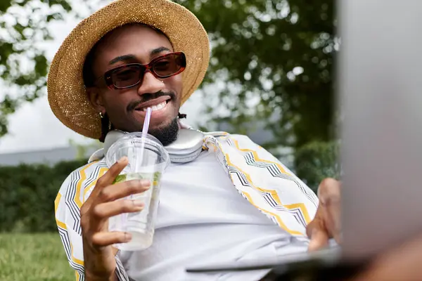 Relaxing in a lively park, a young man works on his laptop while sipping a refreshing drink. — Stock Photo