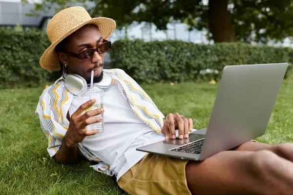 A stylish young man relaxes on the grass, focused on his laptop while sipping a refreshing drink. — Stock Photo