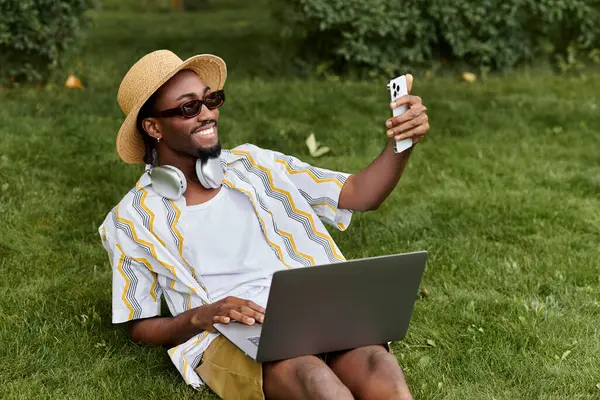 A young african american man relaxes on grass, working on his laptop and taking selfies. — Stock Photo