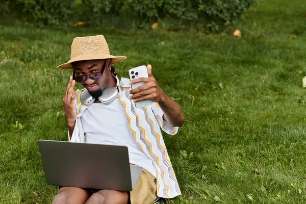 Young man enjoys remote work on his laptop in a vibrant park while using his smartphone. — Stock Photo