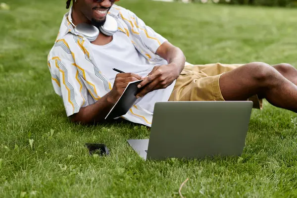 A young african american man relaxes on the grass, jotting down thoughts while using his laptop. — Stock Photo