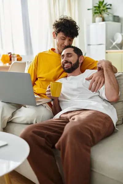 Two men share affection on a sofa while enjoying each others company and a warm drink. — Stock Photo