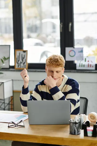 A young redhead man in a striped sweater is intently working at his desk in a bright office. — Stock Photo