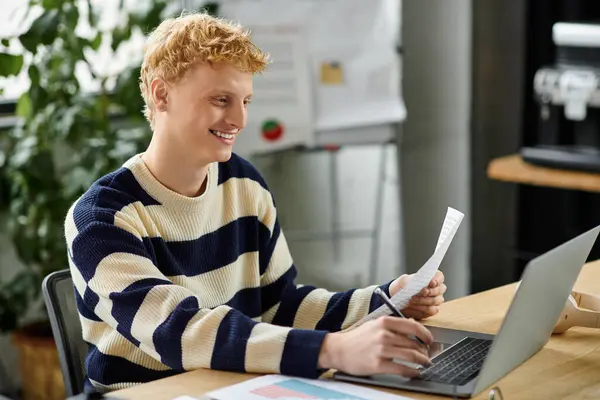 A young redheaded man engages with a laptop while analyzing documents in a bright office. — Stock Photo