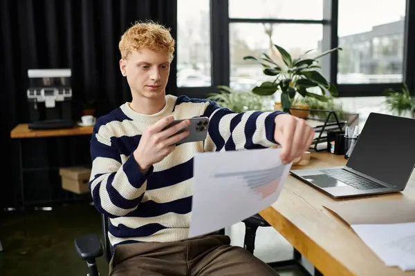 Joven con un suéter a rayas examina los papeles mientras se dedica a su teléfono inteligente en el trabajo. - foto de stock