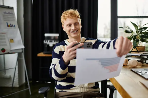 Un joven con el pelo rojo sonríe mientras analiza los informes sobre su teléfono inteligente en el trabajo. - foto de stock