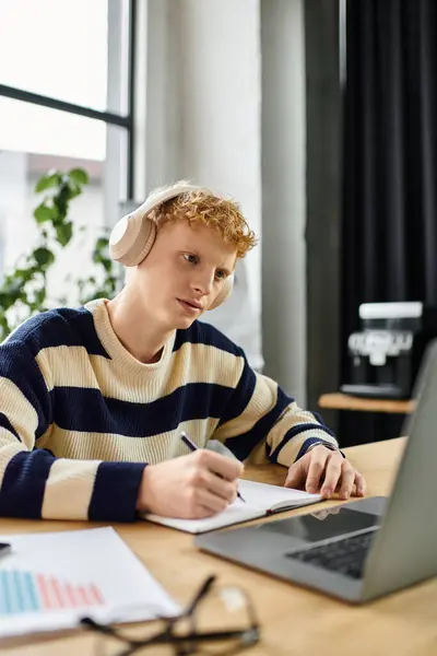 A young redhead man in a striped sweater works diligently at his desk, jotting notes. — Stock Photo