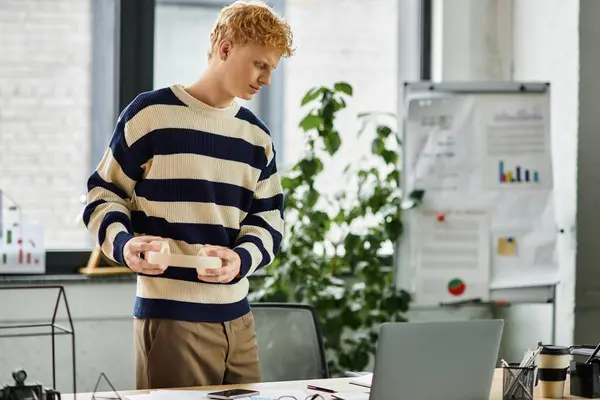 A young redhead man in a striped sweater engages with his work while holding a food container. — Stock Photo