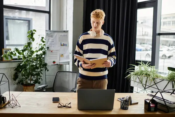 A young man with red hair reviews documents while standing at his desk in a contemporary office. — Stock Photo