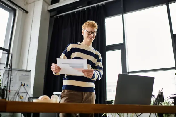 Un jeune homme en pull rayé analyse les rapports tout en se tenant debout dans un espace de travail contemporain. — Photo de stock