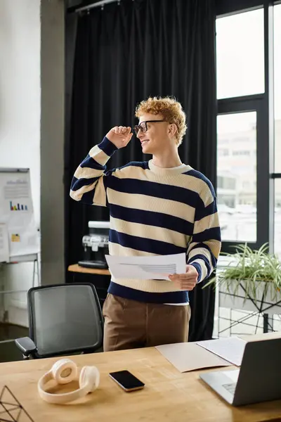 A young redhead man in a striped sweater engages with his team while holding documents at work. — Stock Photo