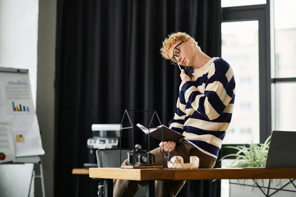 A young redhead man in a striped sweater talks on his phone while sitting stylishly at his desk. — Stock Photo