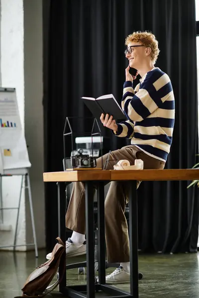 A young redhead man in a striped sweater smiles while reading a book at a modern office desk. — Stock Photo