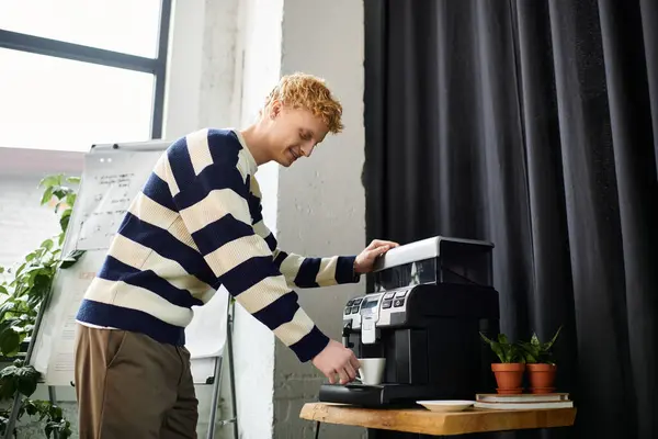 A young redhead man in a striped sweater prepares coffee at his workspace in a corporate office. — Stock Photo