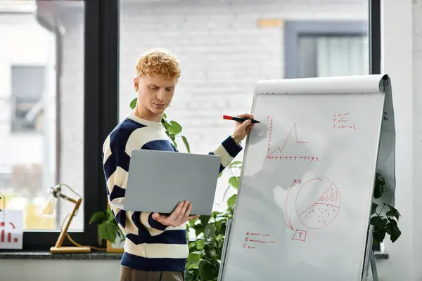 A young man with red hair discusses a chart while using a laptop in a bright office. — Stock Photo