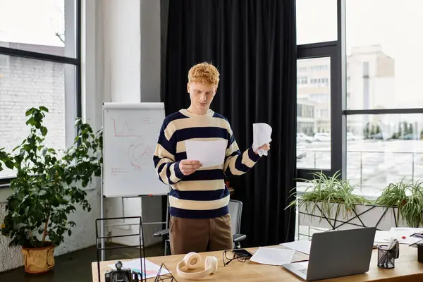 In a contemporary office, a young redhead man reviews documents while standing by his desk. — Stock Photo