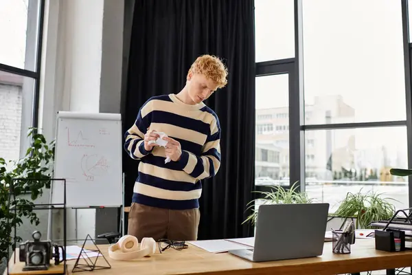 In a stylish office, a young redhead man in a striped sweater reviews notes while working. — Stock Photo