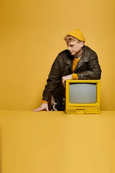 A stylish young man with red hair showcases a trendy winter outfit leaning on a retro TV in a vibrant studio setting. — Stock Photo
