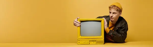 Young man in a trendy winter outfit interacts with a vintage yellow TV against a bright backdrop, banner — Stock Photo
