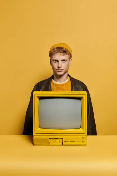 A fashionable young man with red hair showcases his winter attire leaning on a retro TV in a vibrant studio setting. — Stock Photo