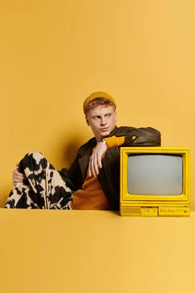 A fashionable young redhead man showcases a chic winter look leaning on a retro TV — Stock Photo