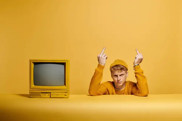 Ein junger rothaariger Mann im Strickkostüm posiert vor leuchtend gelbem Hintergrund in der Nähe von Vintage-Fernsehern und strahlt Stil aus. — Stockfoto