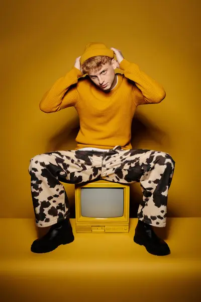 A stylish young man with red hair poses in a trendy winter outfit sitting on vintage tv against a yellow backdrop. — Stock Photo
