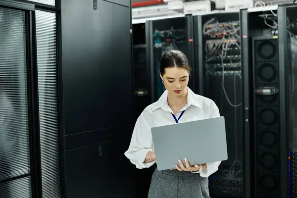 Una donna con una camicia bianca si concentra sul suo computer portatile, supervisionando le operazioni del server in un data center. — Foto stock