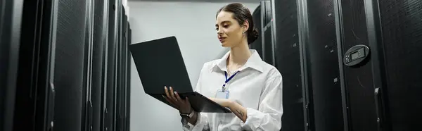 Опытная женщина в белой рубашке работает на ноутбуке посреди серверных стеллажей в дата-центре, баннер — стоковое фото