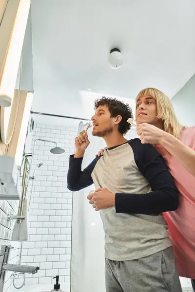 Dos hombres comparten un momento tierno mientras se cepillan los dientes en un baño luminoso y moderno. - foto de stock
